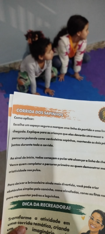 Página da brincadeira “Corrida dos sapinhos” em mãos, com crianças brincando ao fundo.