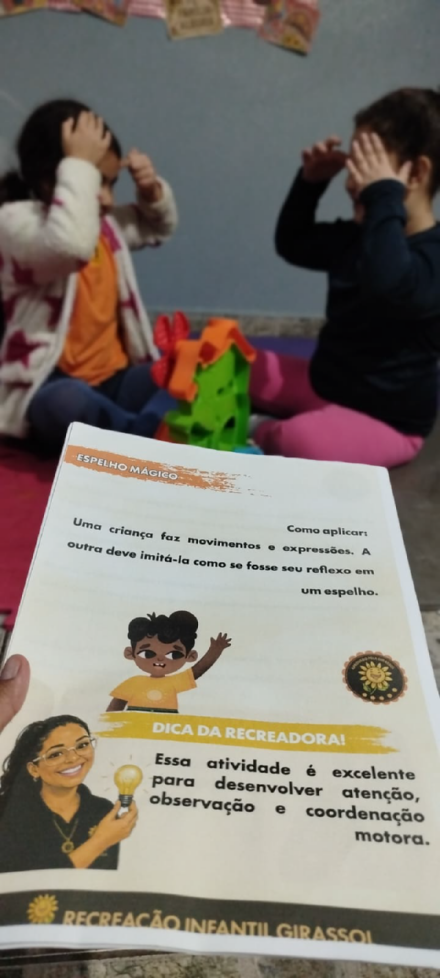 Página da brincadeira “Espelho mágico” em mãos, com crianças imitando gestos ao fundo.